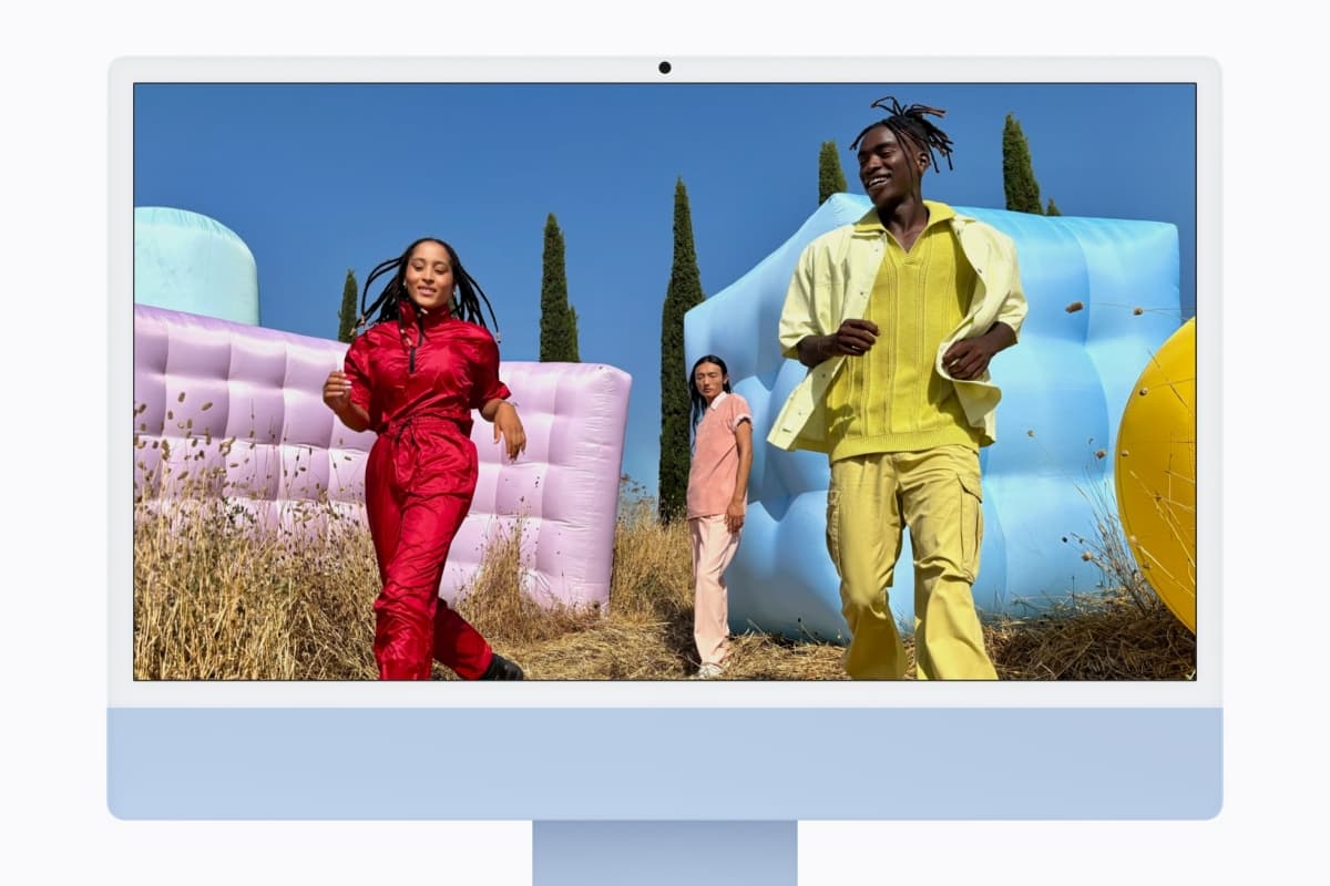 Three people in bright outfits walk past pastel inflatable structures outdoors on a sunny day, with dry grass in the foreground and blue sky behind them.