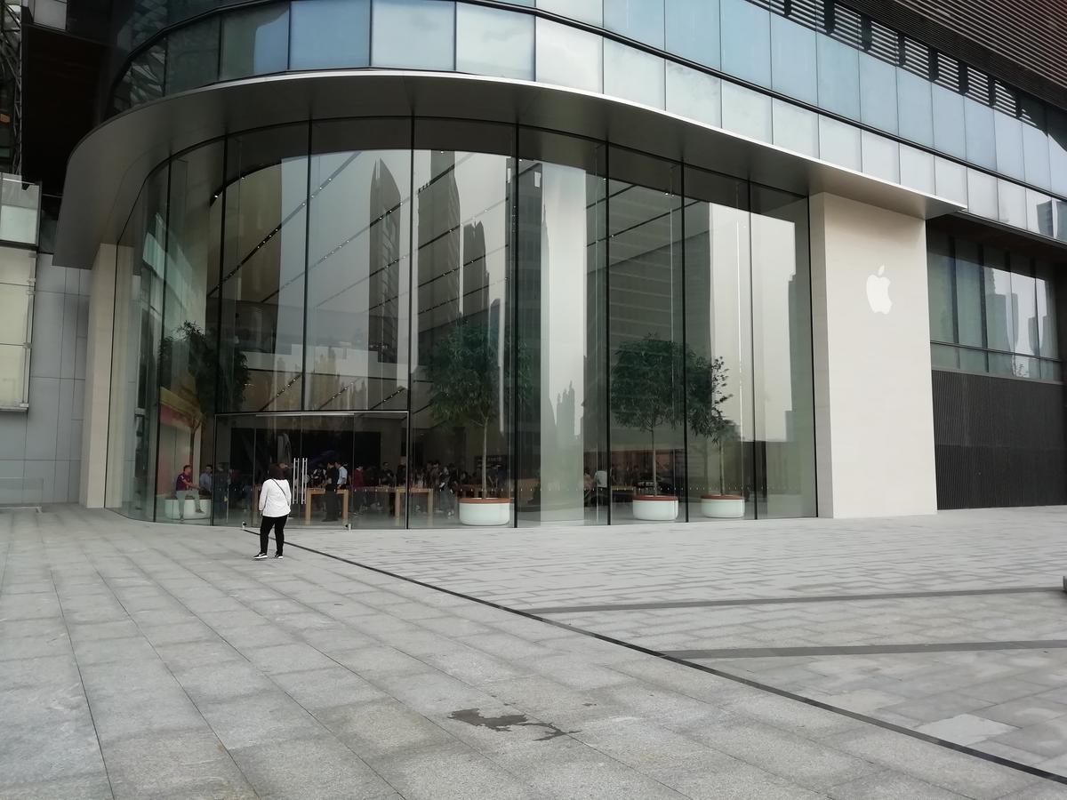 Apple Store in Suzhou Center seen from the plaza, with a curved glass front and the Apple logo on the white wall beside it.