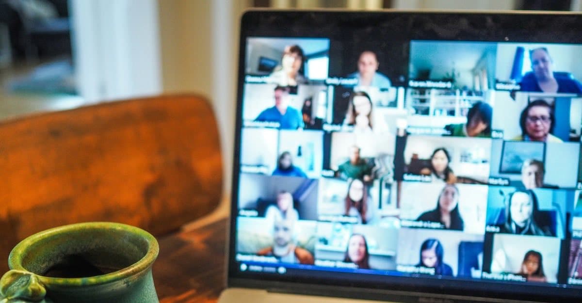 A laptop screen showing a grid of people on a group video call, with a green mug on a wooden table in front