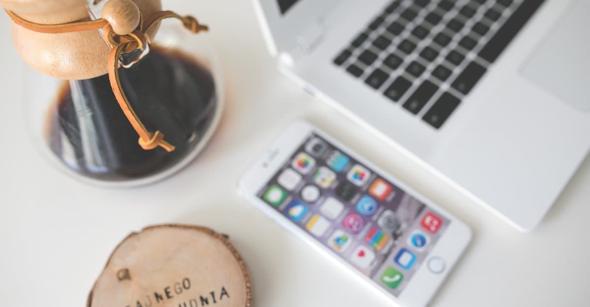 A white iPhone and a silver MacBook on a light desk, beside a glass coffee dripper and a wooden coaster